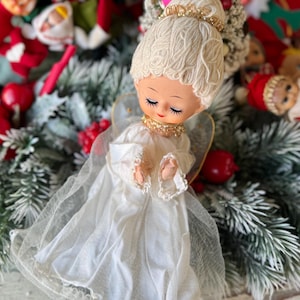 May include: A vintage Christmas angel figurine with a white dress and blonde hair. The angel has closed eyes, a gold collar, and delicate wings. The figurine is surrounded by artificial greenery and red ornaments.