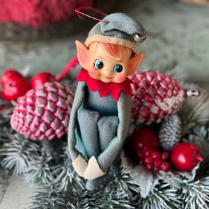 May include: A vintage Christmas elf ornament with a blue felt outfit, red collar, and a blue hat with a silver bell. The elf has rosy cheeks, blue eyes, and orange hair. The ornament is surrounded by festive decorations.