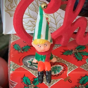 May include: A vintage Christmas elf ornament. The elf has a green and white striped hat, a red scarf, and orange legs with black boots. The ornament is sitting on a red box with a holly pattern. A red "Christmas" sign is in the background.