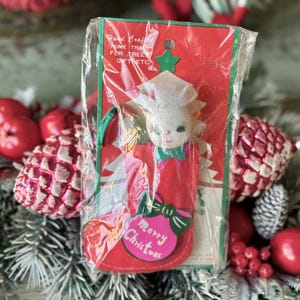 May include: A vintage Christmas ornament in a plastic package. The red felt ornament is shaped like a stocking with a white-faced angel and the words "Merry Christmas". The package reads "Hand Crafted Home Trims for Tree Gift Etc."