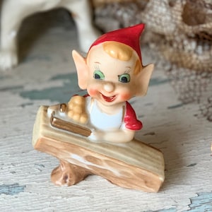 May include: A vintage ceramic figurine of a smiling elf wearing a red hat and sleeves, a white apron, and holding a hammer. The elf is seated on a wooden log. The figurine has a whimsical, antique aesthetic.