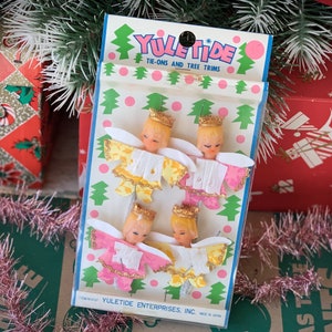 May include: Vintage Yuletide tie-on tree trims featuring four angel ornaments with blonde hair, gold crowns, and colorful wings. The packaging reads "YULETIDE TIE-ONS AND TREE TRIMS" with a pink and green color scheme. The background includes wrapped gifts and tinsel.