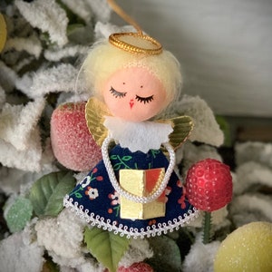 May include: A vintage Christmas angel ornament with a blonde halo and wings. The angel wears a blue dress with a floral pattern and carries a gold gift box. The ornament is surrounded by artificial snow-covered greenery and decorative fruit.