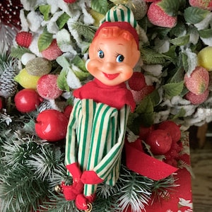 May include: A vintage Christmas elf doll with a smiling face, wearing a red and green striped outfit. The elf is surrounded by a festive arrangement of red apples, frosted berries, and greenery, creating a cheerful holiday scene.