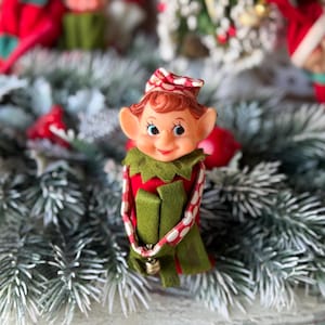 May include: A vintage Christmas elf figurine with a smiling face, wearing a red and green outfit with a bell. The elf has a red and white patterned hat and is surrounded by faux evergreen branches.