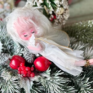 May include: A vintage Christmas angel ornament with a pink face, white hair, and a white dress. The angel is lying on a bed of faux evergreen branches, red berries, and pinecones. A festive holiday decoration.