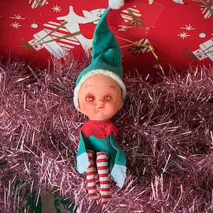 May include: A vintage Christmas elf doll with a green felt hat and outfit, red and white striped legs, and a red knit collar. The doll has a white pom-pom on the hat and is set against a backdrop of pink tinsel and a red box with white and gold designs.