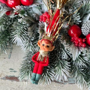 May include: A vintage Christmas elf ornament with a red felt suit, green felt shoes, and a gold-trimmed collar. The elf has a painted face and a red and gold hat. The ornament is surrounded by evergreen branches, red berries, and decorative accents.