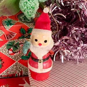May include: A vintage Christmas ornament shaped like Santa Claus. The ornament is red with a white beard and trim, black eyes, and a gold belt buckle. It is set against a festive red and green patterned background.