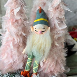 May include: A whimsical elf figurine with a long white beard, rosy cheeks, and a green hat with a yellow band. The elf wears green pants with yellow accents and orange shoes. Two pink feather trees are in the background.