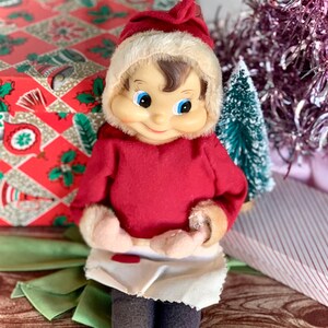 May include: A vintage Christmas elf doll with a red outfit, white apron, and a Santa hat. The doll has a smiling face with blue eyes and brown hair. The background includes a wrapped gift box and a small Christmas tree.