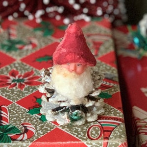 May include: A vintage Christmas ornament featuring a Santa Claus figure. The Santa has a red hat and a white beard, and is attached to a pine cone base. The ornament is set against a festive red and gold patterned background.