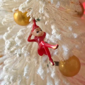 May include: A red plastic elf ornament hanging on a white flocked Christmas tree with gold ornaments.