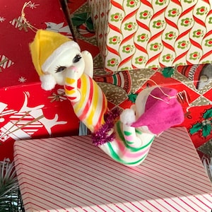 May include: Two vintage Christmas ornaments shaped like candy canes with elf faces. The ornaments are made of plastic and have a yellow, pink, and green striped design. The ornaments are hanging from a Christmas tree.