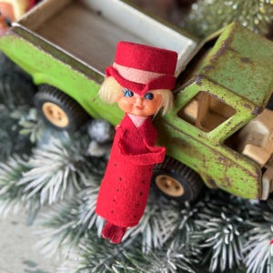 May include: A vintage red doll with a matching hat and coat stands next to a weathered green toy truck. The doll has blonde hair and blue eyes. The truck bed is open, revealing the interior. The scene is set on a bed of faux evergreen branches.