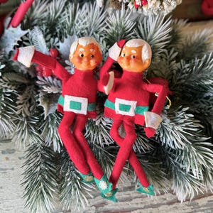 May include: Two vintage Christmas elf ornaments. Each elf is dressed in a red felt suit with a white belt buckle and cuffs, green accents, and green shoes. The elves have white hair and smiling faces. They are posed on a bed of evergreen branches.