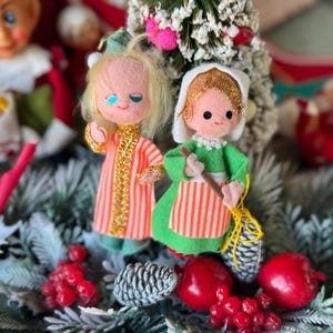 May include: Two vintage Christmas ornaments. One doll wears a pink and orange striped robe with gold trim. The other doll wears a green dress with an orange and white striped apron and holds a broom. The ornaments are surrounded by greenery and red berries.