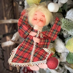 May include: A vintage Christmas angel ornament with a plaid red and green fabric dress. The angel has a blonde head and closed eyes, with a white lace trim. The ornament is surrounded by artificial greenery and fruit.