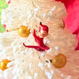 May include: A small, white Christmas tree with a flocked appearance, adorned with gold ornaments and a red elf figurine. The elf is in a climbing pose, adding a playful touch to the festive decor. The background is a vibrant red.