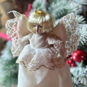 May include: A porcelain angel tree topper with a cream-colored dress and blonde hair. The angel has lace-trimmed wings and a gold halo. The dress is accented with gold trim. The angel is a Christmas decoration.