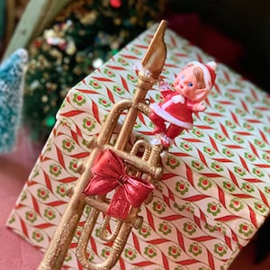 May include: A gold-colored trumpet Christmas ornament with a red bow and a small elf figure in a red and white outfit. The ornament is placed in front of a patterned gift box with red and green floral designs.
