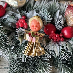 May include: A close-up of a Christmas tree decoration featuring a vintage angel figurine with a gold foil dress and a blonde head. The angel is surrounded by red berries, apples, and frosted pine needles, creating a festive holiday scene.