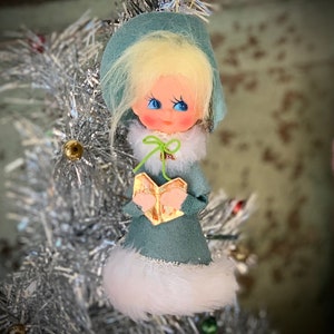 May include: A vintage Christmas ornament featuring a doll dressed in a teal-colored outfit with a white fur trim. The doll has blonde hair, rosy cheeks, and is holding a small, golden book. The ornament is displayed on a silver tinsel Christmas tree.