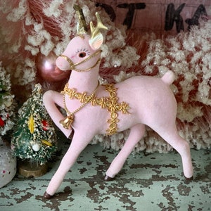 May include: A pink, vintage-style deer figurine with gold-colored antlers and decorative accents. The deer wears a gold-colored harness and a bell. The figurine is set against a backdrop of a pink Christmas tree and other festive decorations.