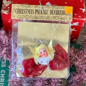 May include: Vintage Christmas package decorations featuring an angel with blonde hair, a red dress, and gold wings. The package is sealed in plastic and includes the text "CHRISTMAS PACKAGE DECORATIONS".