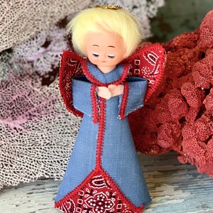 May include: A handcrafted angel figurine with a denim-like blue dress and red bandana-patterned wings and accents. The angel has blonde hair, closed eyes, and a gold halo. The figurine is a decorative item.
