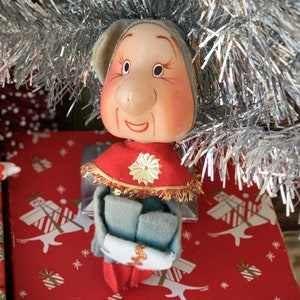 May include: A vintage Christmas elf figurine with a painted face, wearing a red cape with gold trim and a matching hat. The elf is holding a scroll and is sitting on a red gift box with a white gift pattern. Silver tinsel is in the background.