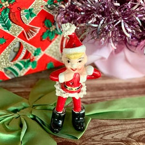 May include: A vintage Christmas elf figurine in a red Santa suit with black boots and a white trim. The elf is standing on a green satin bow, with a red and gold patterned gift box in the background.