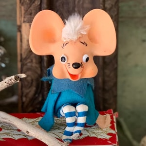 May include: A vintage mouse doll with large ears, a white tuft of hair, and a blue felt outfit. The doll has striped blue and white legs and is sitting on a red and gold patterned surface. The mouse has a cheerful expression.