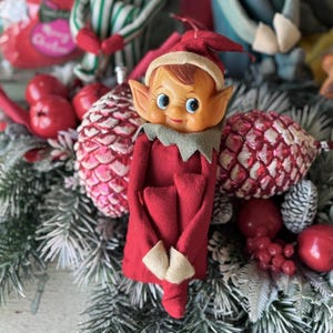 May include: A vintage Christmas elf doll with a red felt outfit, a smiling face, and a pointed hat. The elf is surrounded by festive decorations, including red ornaments, pinecones, and greenery, creating a holiday scene.