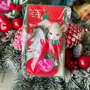 May include: A vintage Christmas ornament featuring a white mouse head emerging from a red stocking. The ornament is packaged in a clear plastic sleeve with a red and green cardboard backing. The backing reads "Merry Christmas".