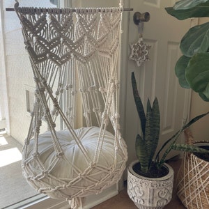 May include: Cream-colored macrame hanging chair with a round, off-white cushion. The chair is suspended from a metal bar and features intricate woven patterns. A decorative star hangs on the door behind the chair. A potted snake plant and a woven basket are also visible.