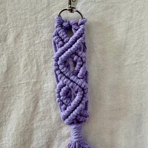 May include: A handmade macrame keychain in a soft lavender color. The keychain features a woven design with a small tassel at the bottom and a silver-colored clasp at the top for easy attachment to keys or bags.