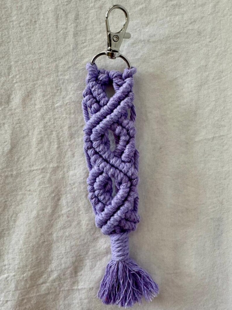 May include: A handmade macrame keychain in a soft lavender color. The keychain features a woven design with a small tassel at the bottom and a silver-colored clasp at the top for easy attachment to keys or bags.