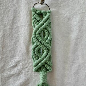 May include: A handmade macrame keychain in a light green color. The keychain features a woven design with a tassel at the bottom and a silver-colored clasp. The macrame is made of a soft, textured material.