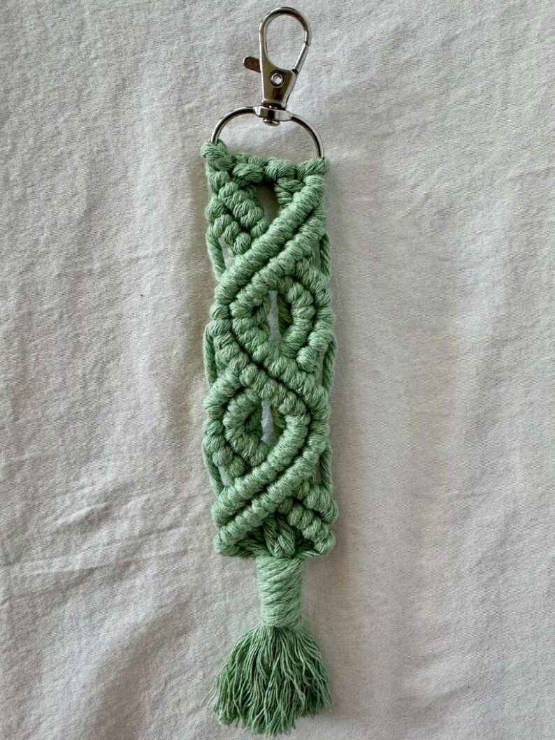 May include: A handmade macrame keychain in a light green color. The keychain features a woven design with a tassel at the bottom and a silver-colored clasp. The macrame is made of a soft, textured material.