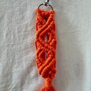 May include: Bright orange macrame keychain with a silver-colored clasp. The keychain features a woven design with a tassel at the bottom. The macrame is made of a thick, durable cord.