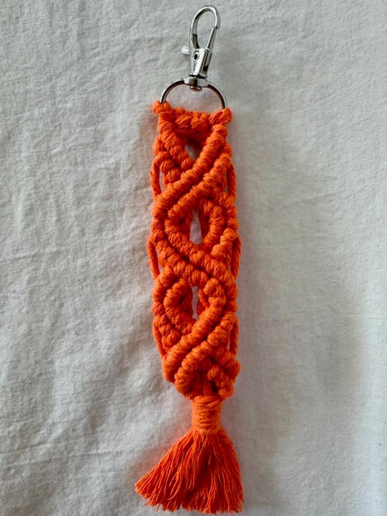 May include: Bright orange macrame keychain with a silver-colored clasp. The keychain features a woven design with a tassel at the bottom. The macrame is made of a thick, durable cord.