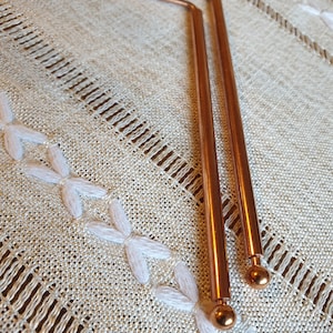 May include: Two copper-colored metal rods with rounded ends. The rods are bent to form a right angle at the top and are lying on a white fabric surface.