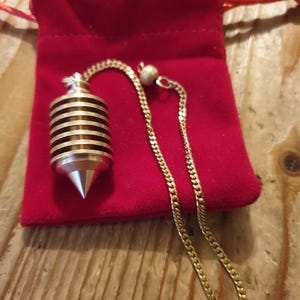 Può includere: Un pendolo con una catena d'oro e un sacchetto di velluto rosso. Il pendolo ha una punta argentata e un corpo cilindrico con bande dorate e marroni alternate. La catena è una catena a maglia d'oro.