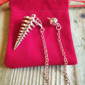 Könnte beinhalten: Ein rosegoldfarbenes Metallpendel mit spiralförmigem, kegelförmigem Design, das an einer feinen Gliederkette befestigt ist. Die Kette weist nahe dem oberen Ende eine kleine, kugelförmige Filigranperle auf und liegt auf einem roten Samtbeutel.