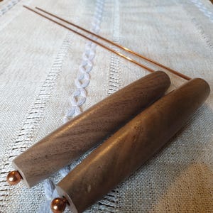 Op de afbeelding: Twee houten deuvels met koperen accenten en twee koperen staven liggen op een wit linnen oppervlak. De deuvels zijn cilindrisch en hebben een donkerbruine afwerking. De koperen staven zijn dun en recht.