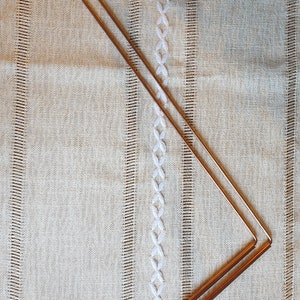 Può includere: Due aste metalliche dorate collegate ad angolo retto, formando un angolo di 90 gradi. Le aste sono appoggiate su una superficie di tessuto beige con un motivo ricamato bianco.
