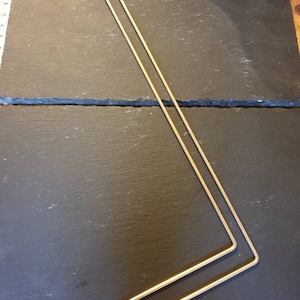 May include: Two thin, gold-colored metal rods, bent at a right angle, are positioned on a dark, textured surface. The rods are parallel to each other, creating a geometric design. The background is a dark, possibly slate-like material.