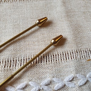 May include: Two gold-coloured metal tools with pointed tips, on a beige linen surface with white embroidered floral details. The tools have a slender, rod-like body and a conical tip.