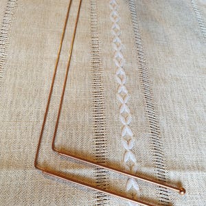 May include: Two long, thin, copper knitting needles with rounded tips. The needles are lying on a white, textured fabric.
